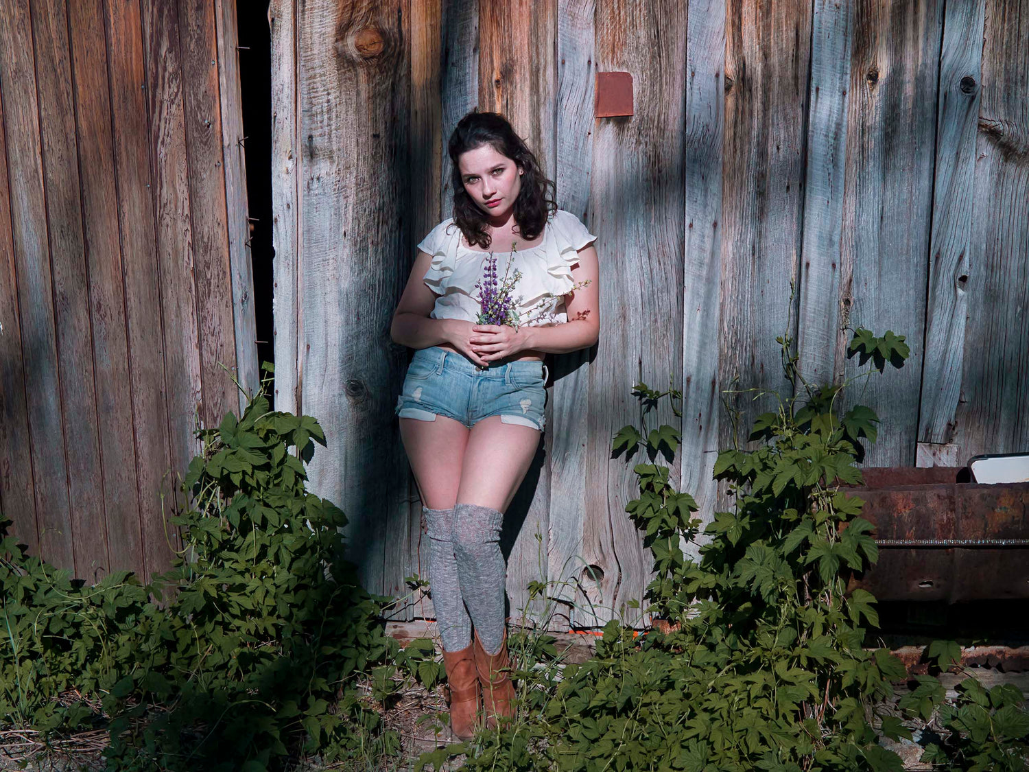 Xan stands in front of old textured barn wood, near a patina metal trough. She wears a delicate white blouse, denim shorts, browinsh grey stockings, brown boots and holds wild purple flowers in her hands. She is framed by wild green climbing ivy. 