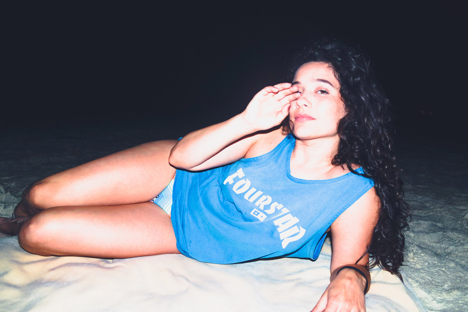 Xan reclines on a yellow towel on the beach at night wearing a bright blue tank top, her long curly dark hair cascades over her shoulder. 