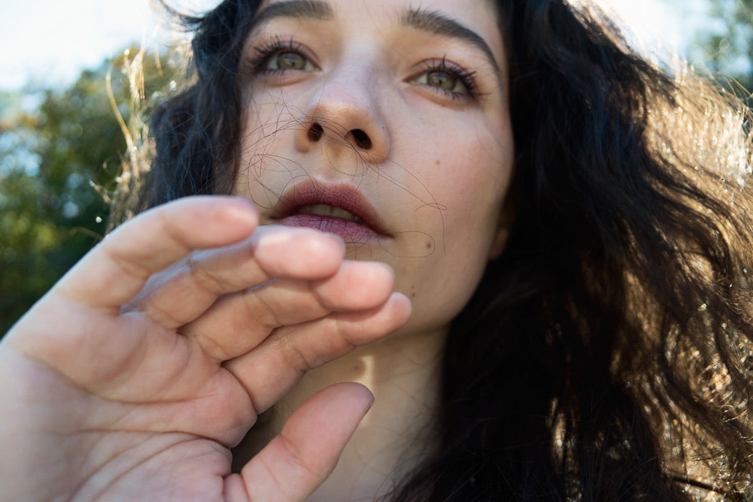 In a close up portrait Xan stands backlit in front of the camera with green eyes, berry colored lips, and sunlight peeking through her dark curly hair. 