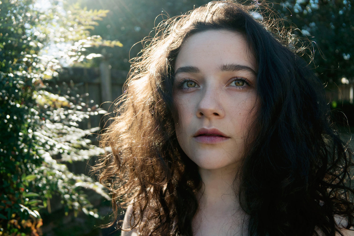 In a close up portrait Xan stands backlit in front of the camera with green eyes, berry colored lips, and sunlight peeking through her dark curly hair. 