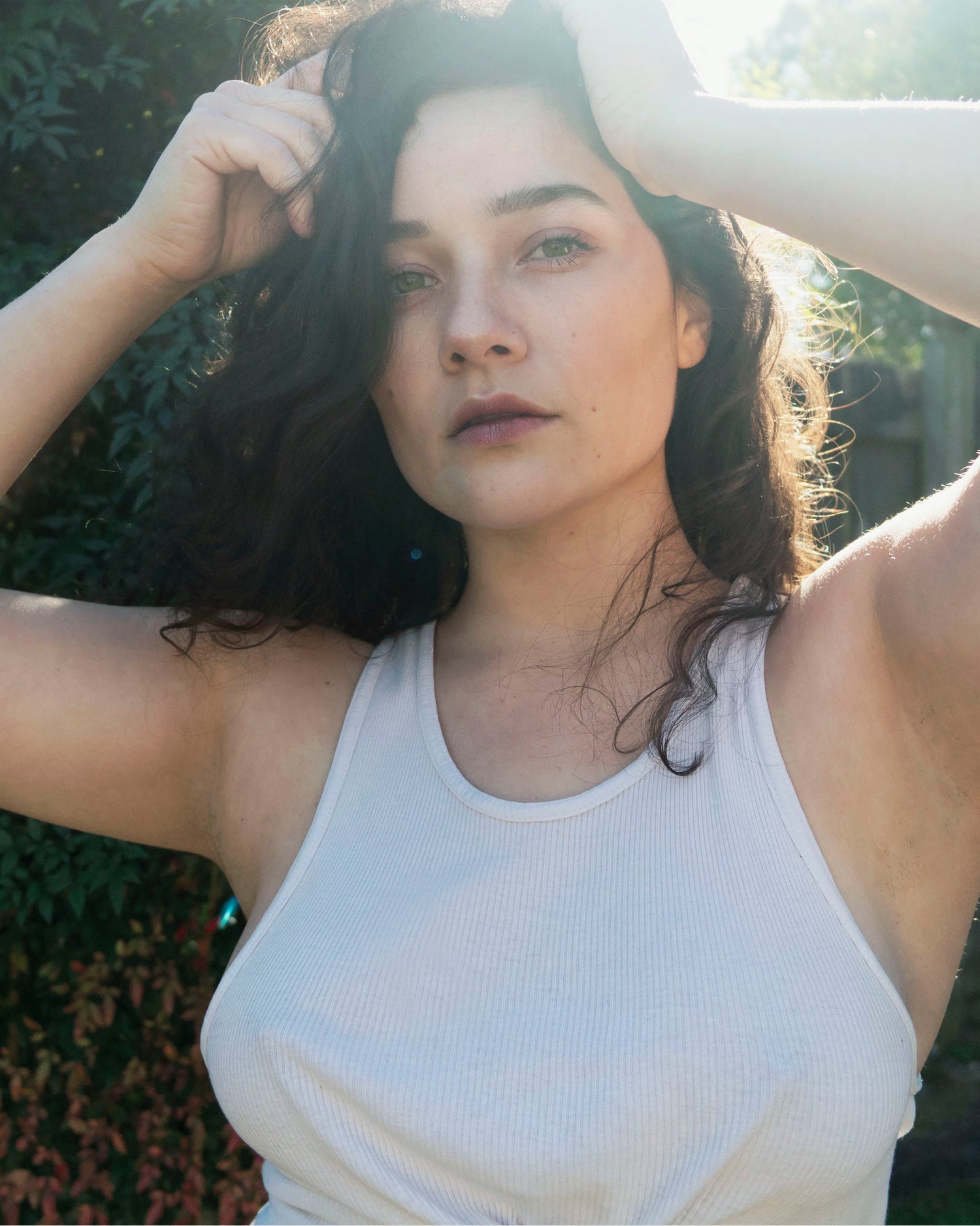 In a medium portrait Xan stands backlit in front of the camera with green eyes, berry colored lips, and sunlight peeking through her dark curly hair while wearing a white tank top. . 