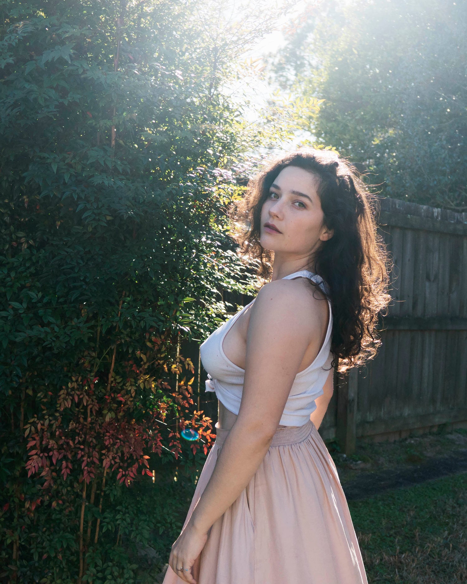 In a close up portrait Xan stands backlit in front of the camera with green eyes, berry colored lips, and sunlight peeking through her dark curly hair in a pale pink skirt and white tank top in front of green bushes.