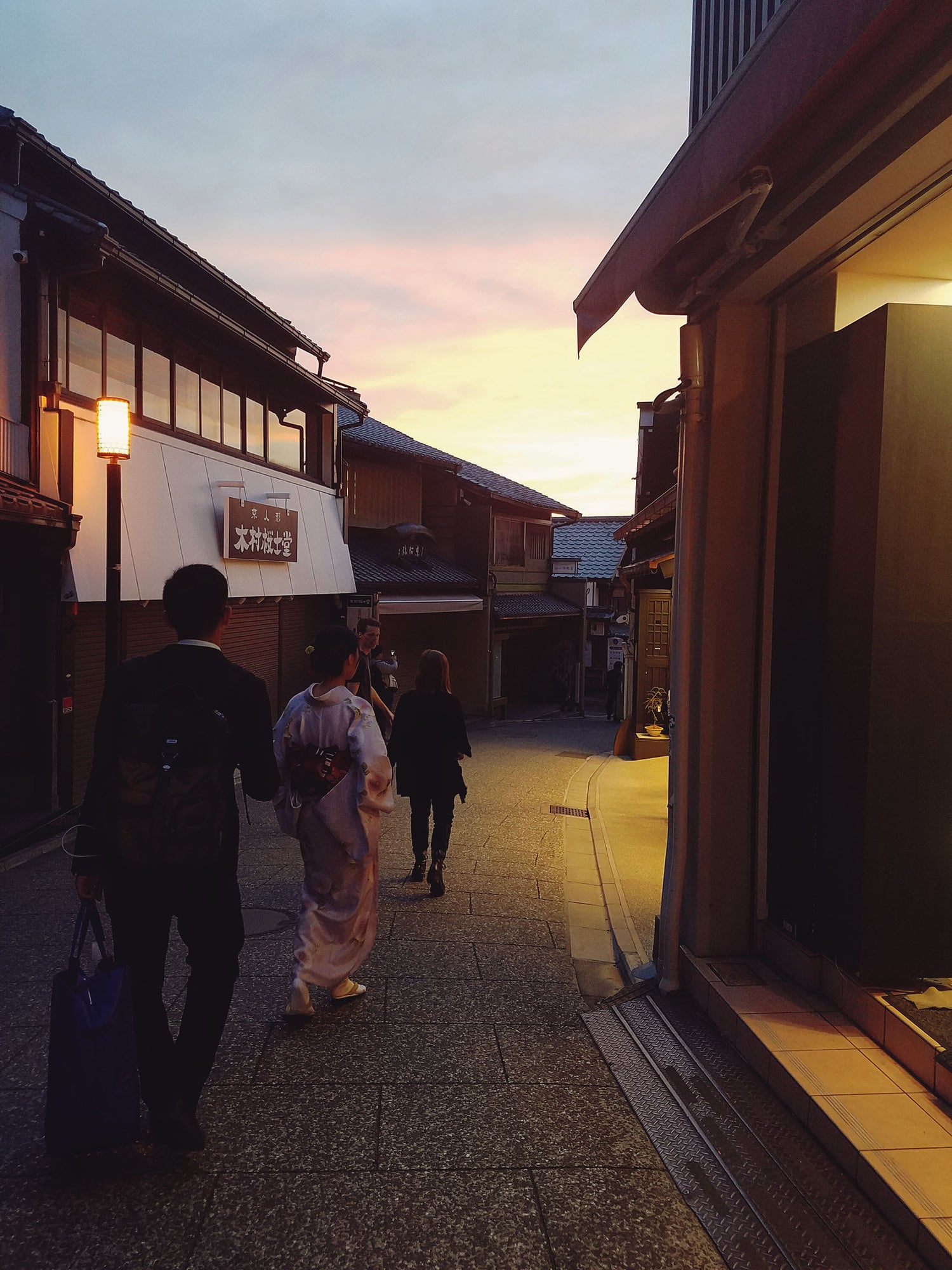 Sunset on the Gion strip in Kyoto Japan during Cherry Blossom festivities around the city. An elegant woman in a pastel pink Kimono walks down the dimply lit cobbled street towards the sunset. 