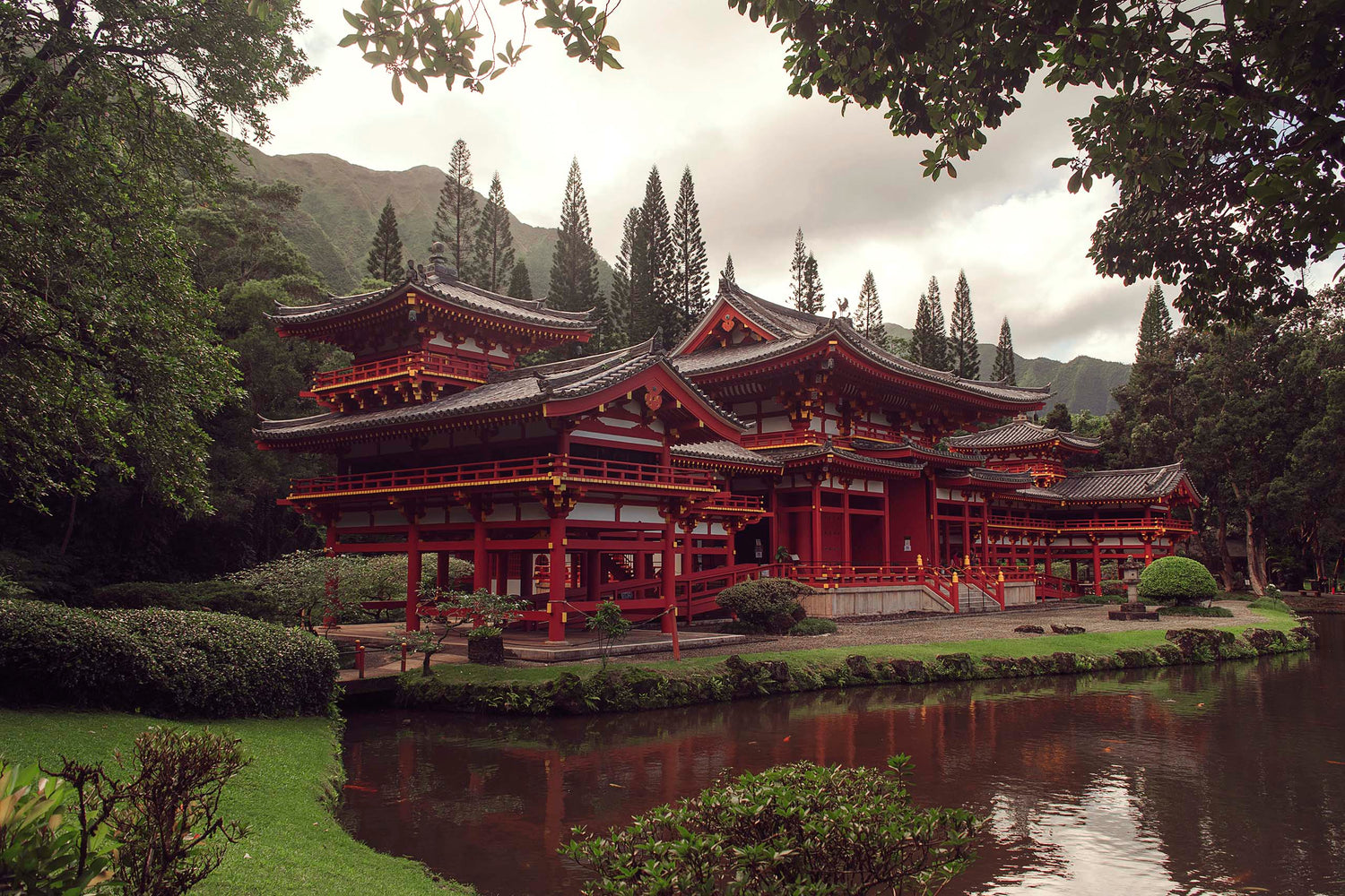 The magnificent Byodo In Temple in Honolulu, Hawaii framed by the magnificent grounds surrounding the temple. The brilliant red temple stands proudly amidst the green foliage of the garden and the jungled mountains behind.