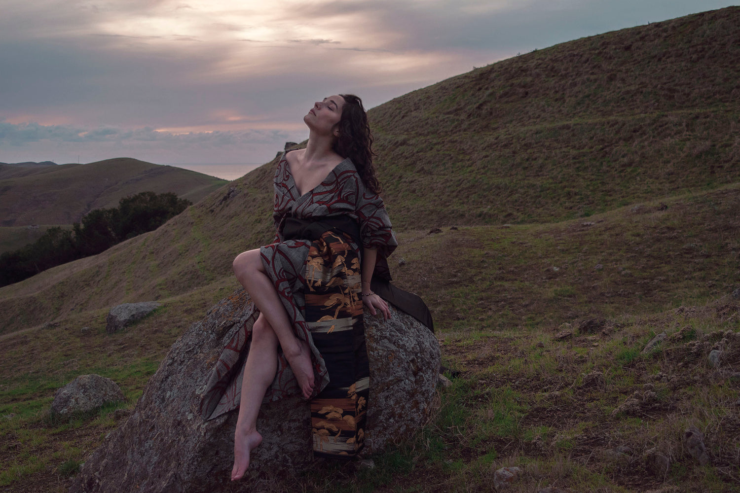 Xan from Stories of Evermore sits on a large orange moss covered grey stone in a red and grey kimono with a black, gold, copper, and silver obi belt tie amidst rolling hills at sunset.