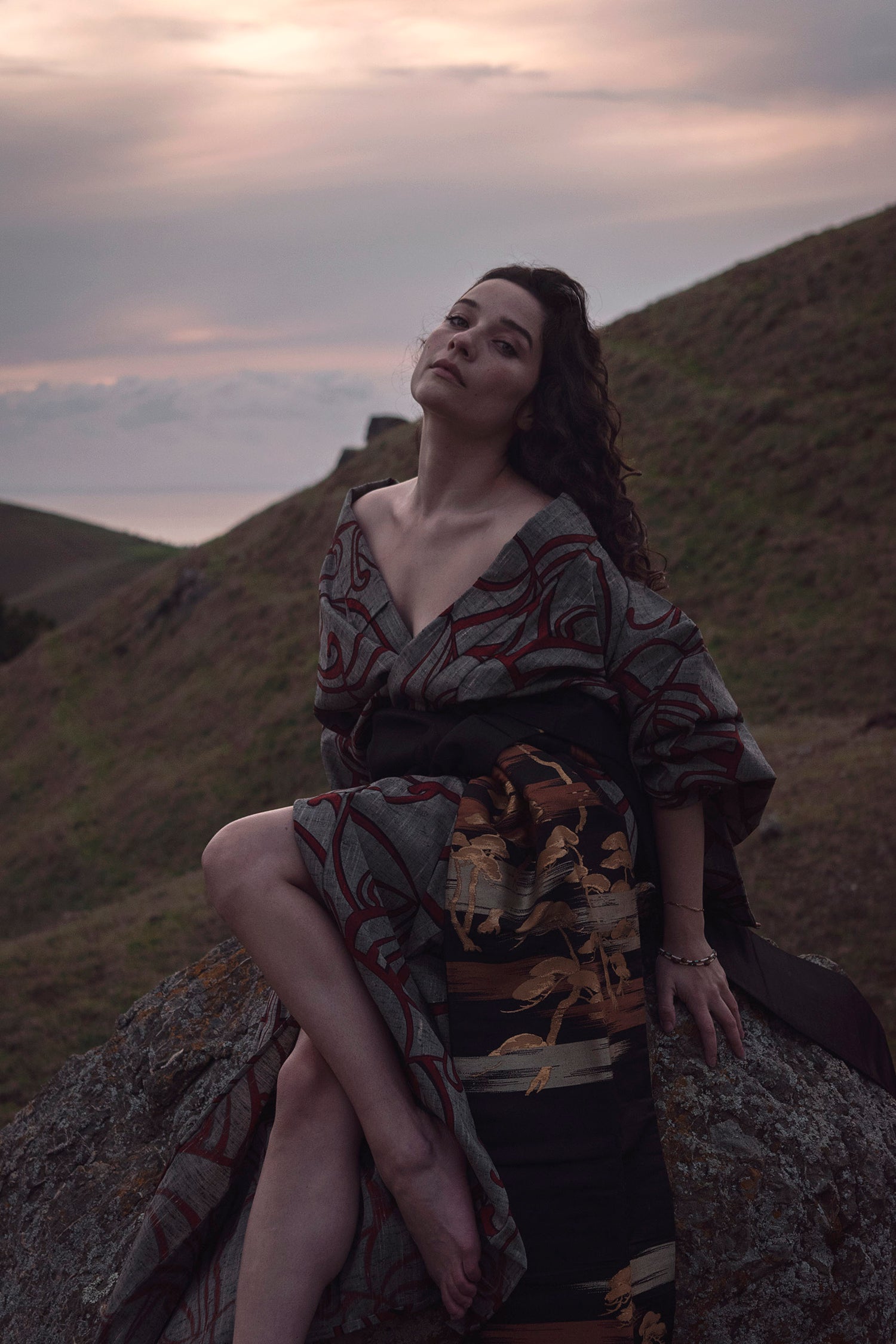 Xan from stories of evermore sits on a large orange moss covered grey stone in a red and grey kimono with a black, gold, copper, and silver obi belt tie amidst rolling hills at sunset.