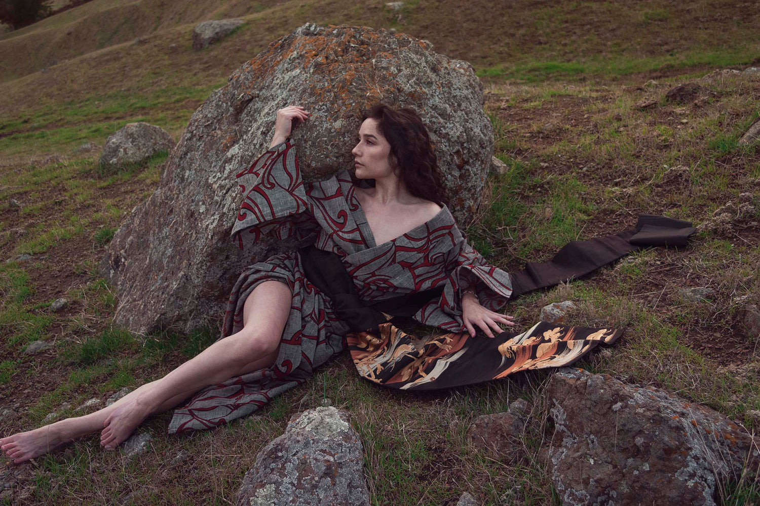 Xan from stories of evermore lays in front of a large orange moss covered grey stone in a red and grey kimono with a black, gold, copper, and silver obi belt tie amidst the grass.