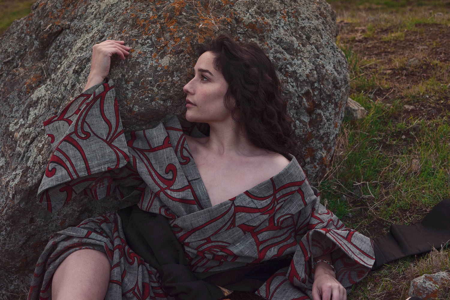 Xan from stories of evermore lays in front of a large orange moss covered grey stone in a red and grey kimono with a black, gold, copper, and silver obi belt tie amidst the grass.