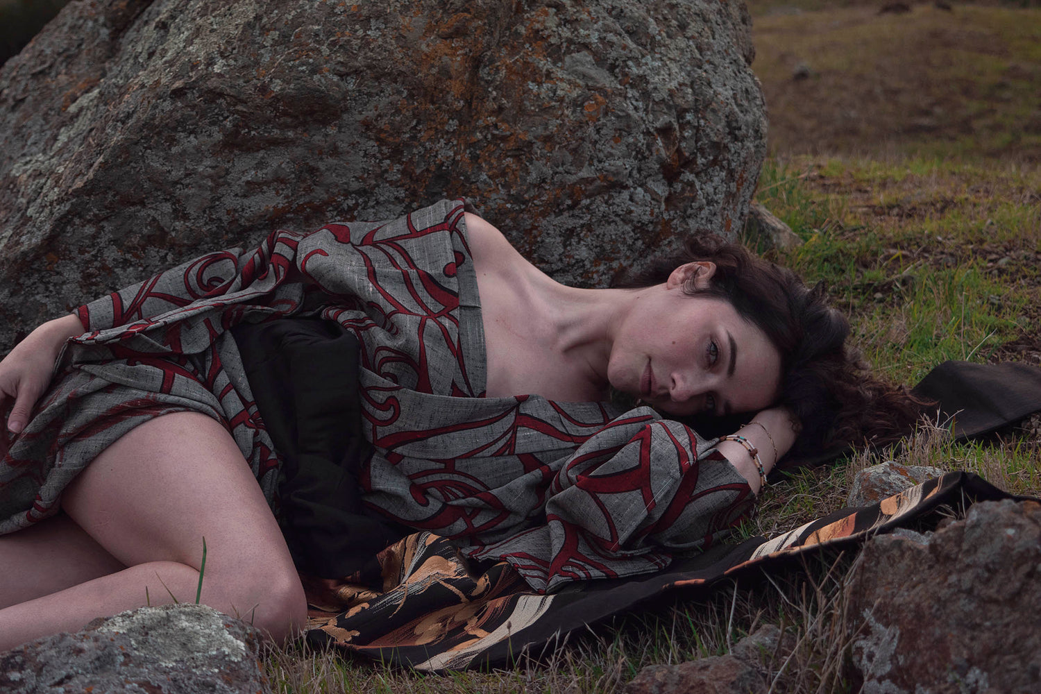 Xan from stories of evermore lays in front of a large orange moss covered grey stone in a red and grey kimono with a black, gold, copper, and silver obi belt tie amidst the grass.