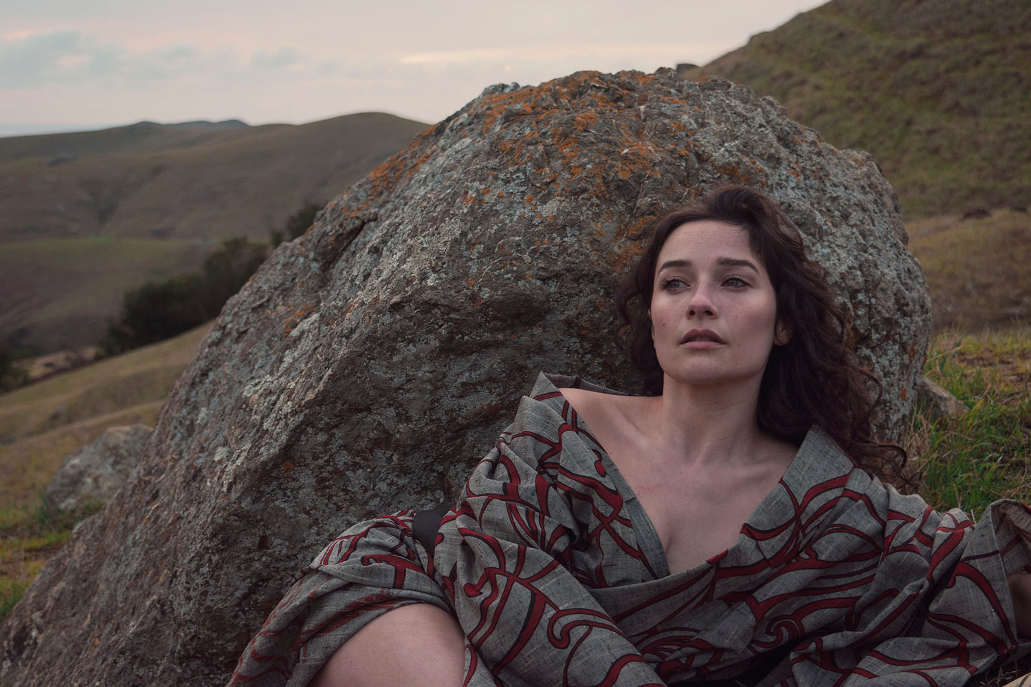 Xan from stories of evermore lays in front of a large orange moss covered grey stone in a red and grey kimono with a black, gold, copper, and silver obi belt tie amidst the grass.