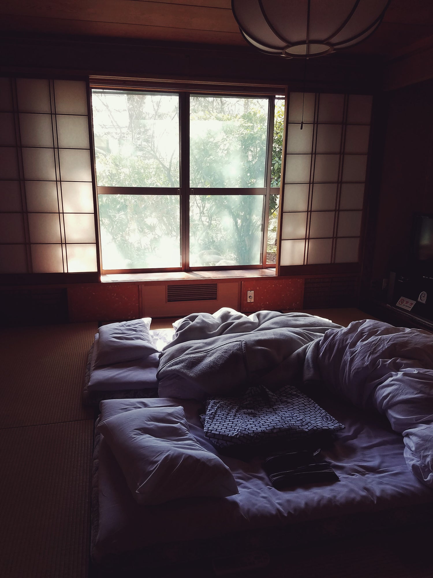 Japanese futon style sleeping arrangements in an Onsen (Japanese Hot Spring Inn) bathed in sunlight framed by shoji screens, atop a tatami mat. 