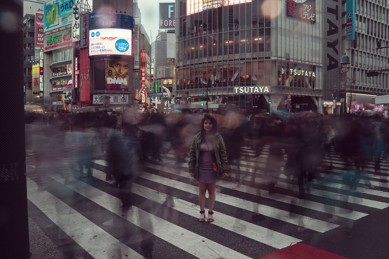 Xan from Stories of Evermore is standing in the middle of the cross walk in Shibuya Tokyo. She stands still amidst a ghostly sea of people. The bright lights of the stores shine from the skyscrapers behind her. 