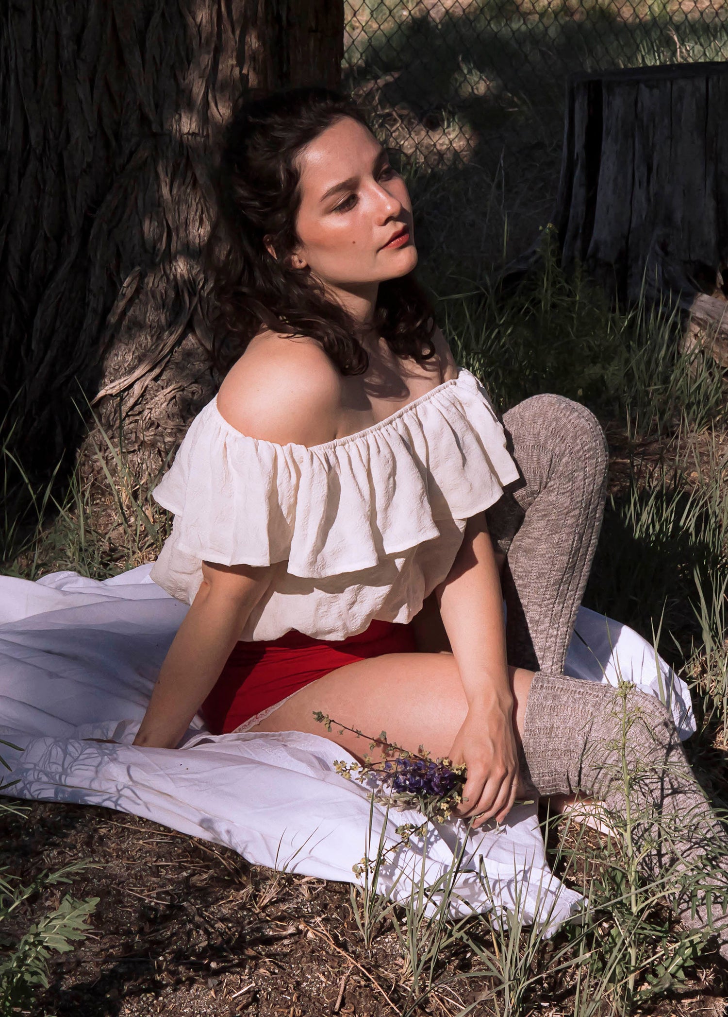 A romantic portrait of Xan underneath an abandoned tree house in a ruffled white blouse, red undergarments, in brownish thigh high stockings. She holds purple wild flowers in her hands and sits on a white blanket. 