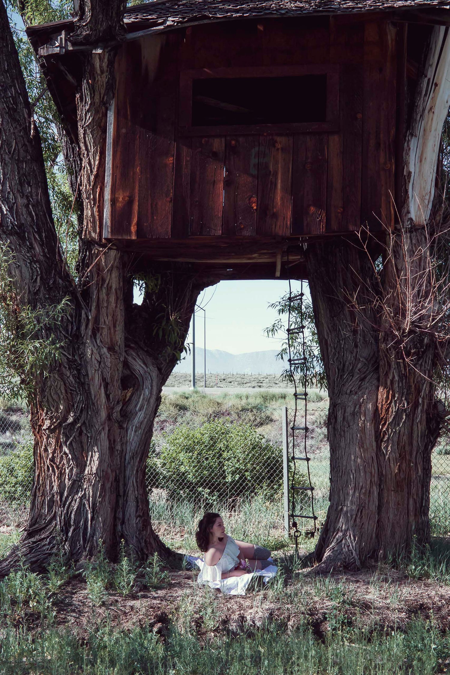 A romantic portrait of Xan underneath an abandoned tree house in a ruffled white blouse, red undergarments, in brownish thigh high stockings. She holds purple wild flowers in her hands and sits on a white blanket. 