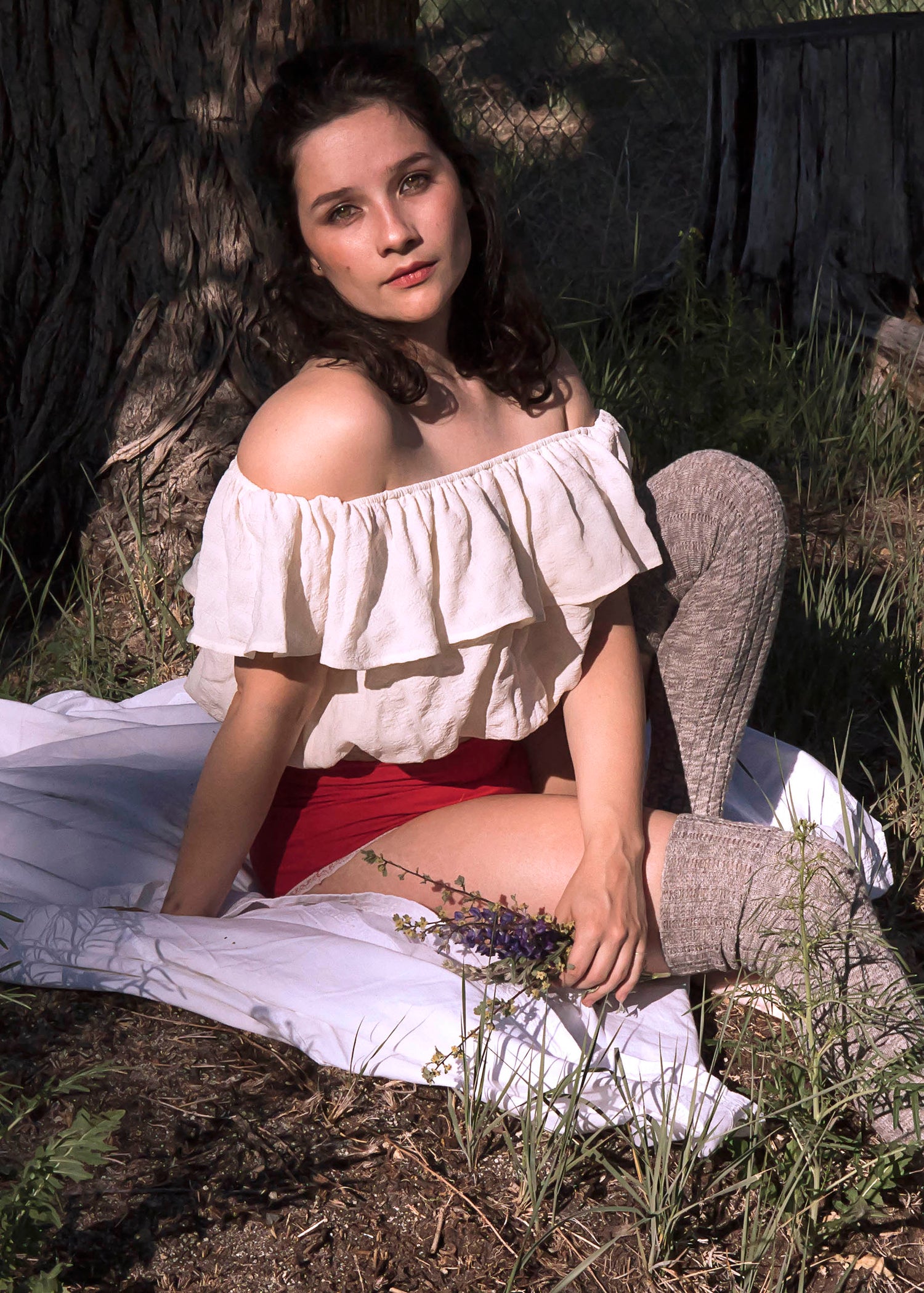 A romantic portrait of Xan underneath an abandoned tree house in a ruffled white blouse, red undergarments, in brownish thigh high stockings. She holds purple wild flowers in her hands and sits on a white blanket. 