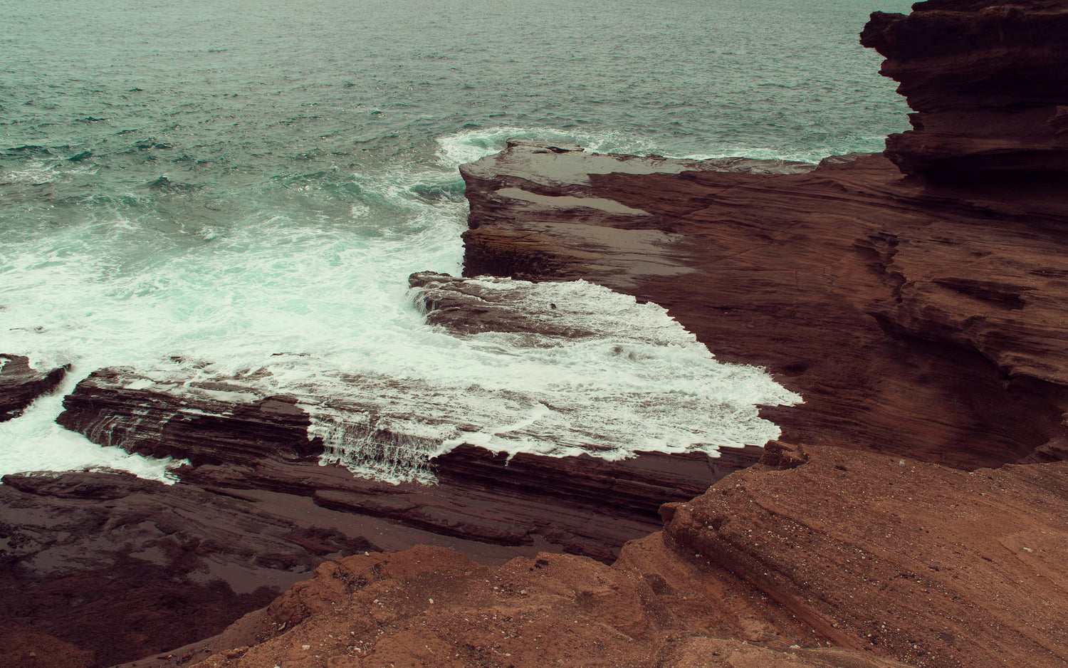 The Pacific Ocean gently caresses the brown and black volcanic rock in the sea green waters of Honolulu, Hawaii.