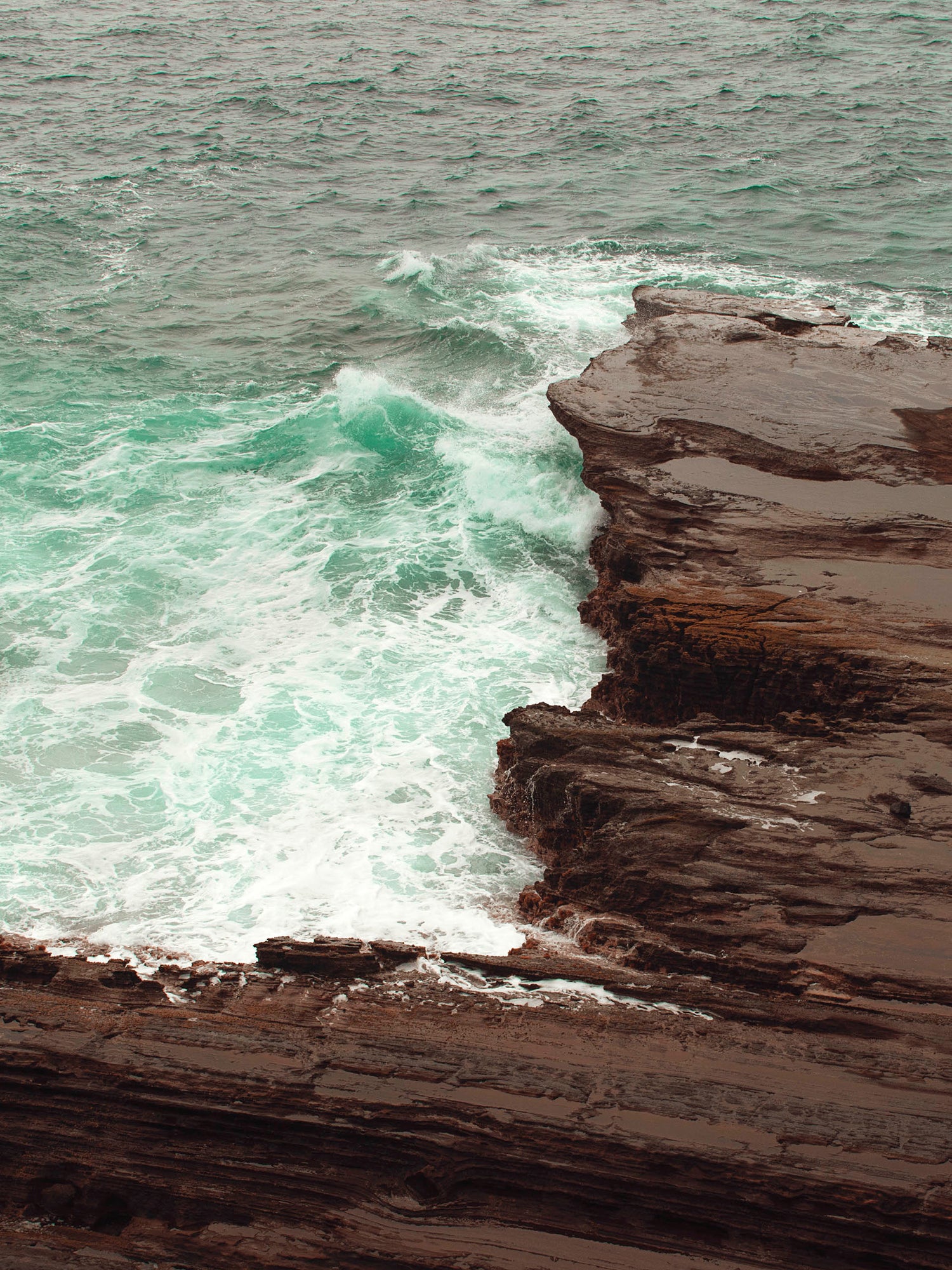 The Pacific Ocean gently caresses the brown and black volcanic rock in the sea green waters of Honolulu, Hawaii.