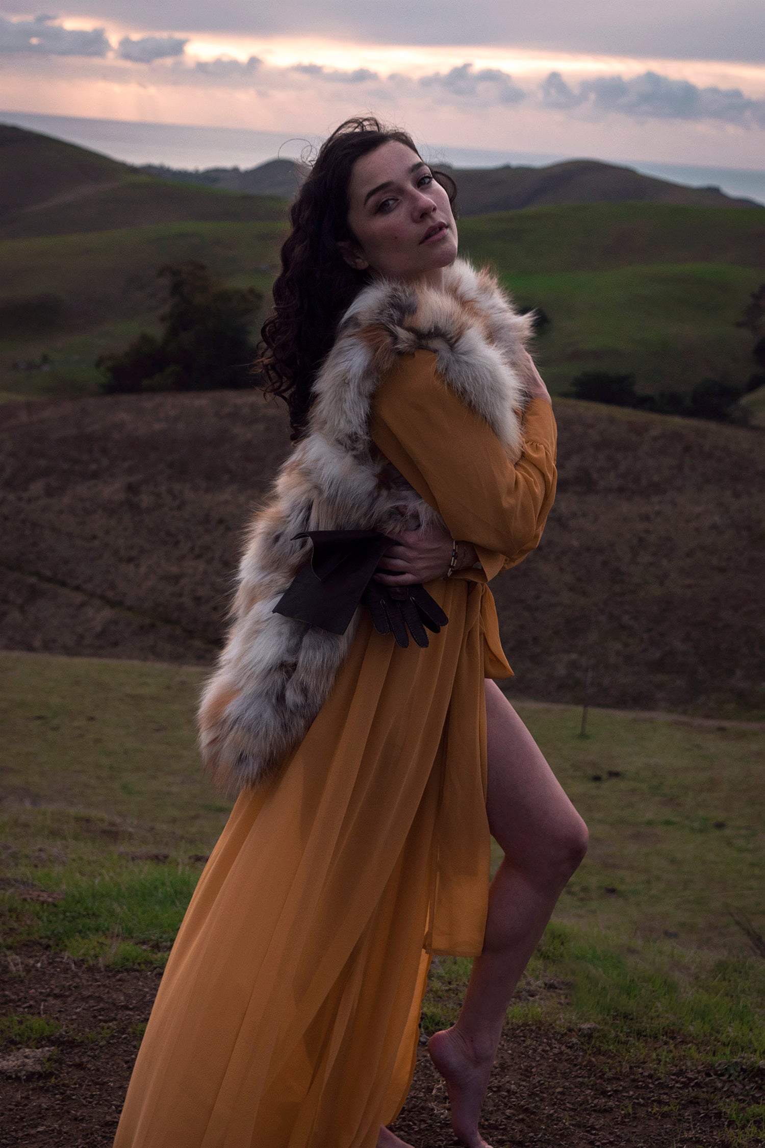 Xan from Stories of Evermore stands in front of a sprawling vista of green rolling hills and the distant ocean at sunset in a mustard yellow gown and vintage fur vest and while holding vintage leather gloves.