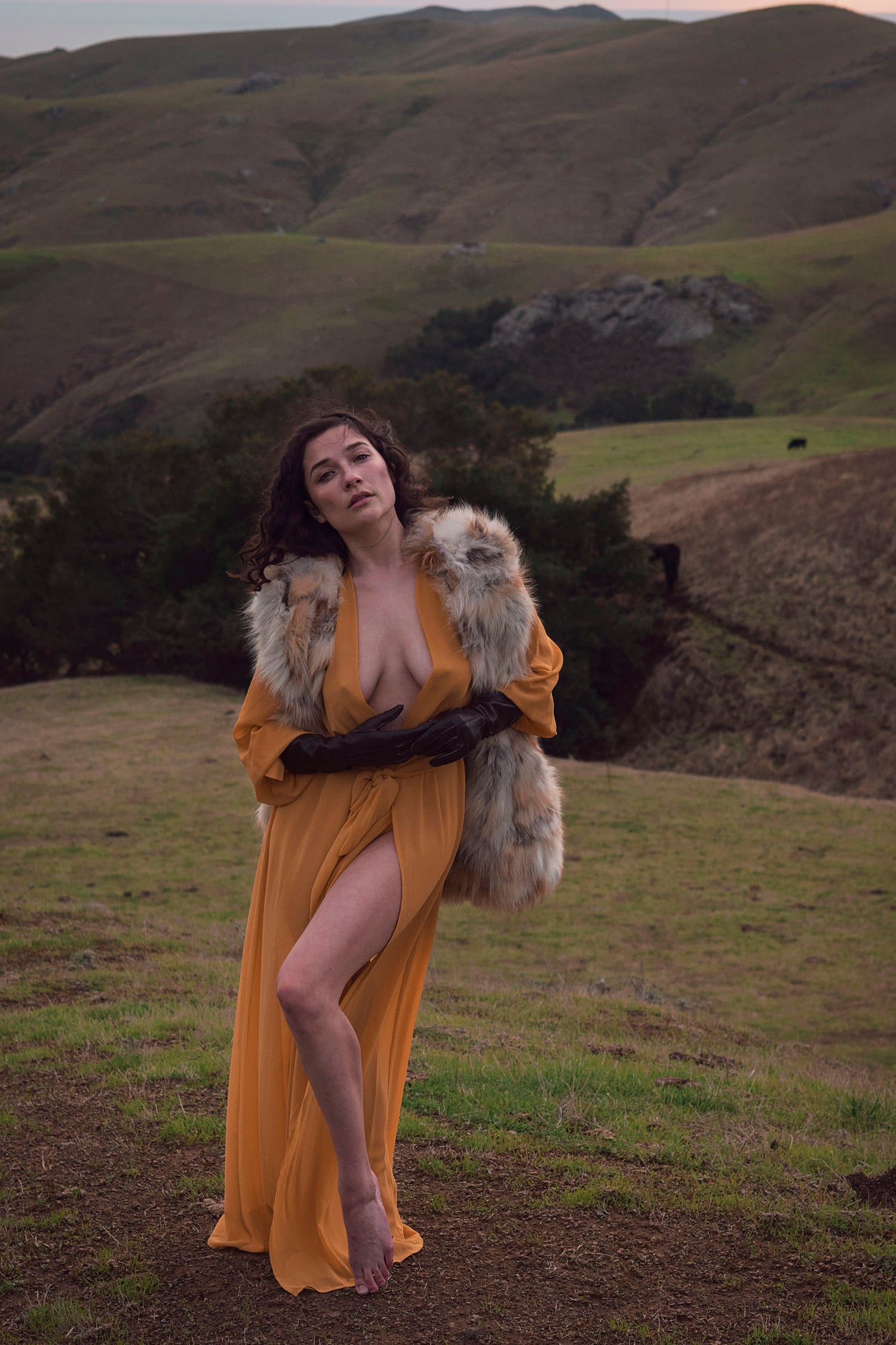 Xan from Stories of Evermore stands in front of a sprawling vista of green rolling hills and the distant ocean at sunset in a mustard yellow gown while wearing a vintage fur vest and vintage leather gloves.