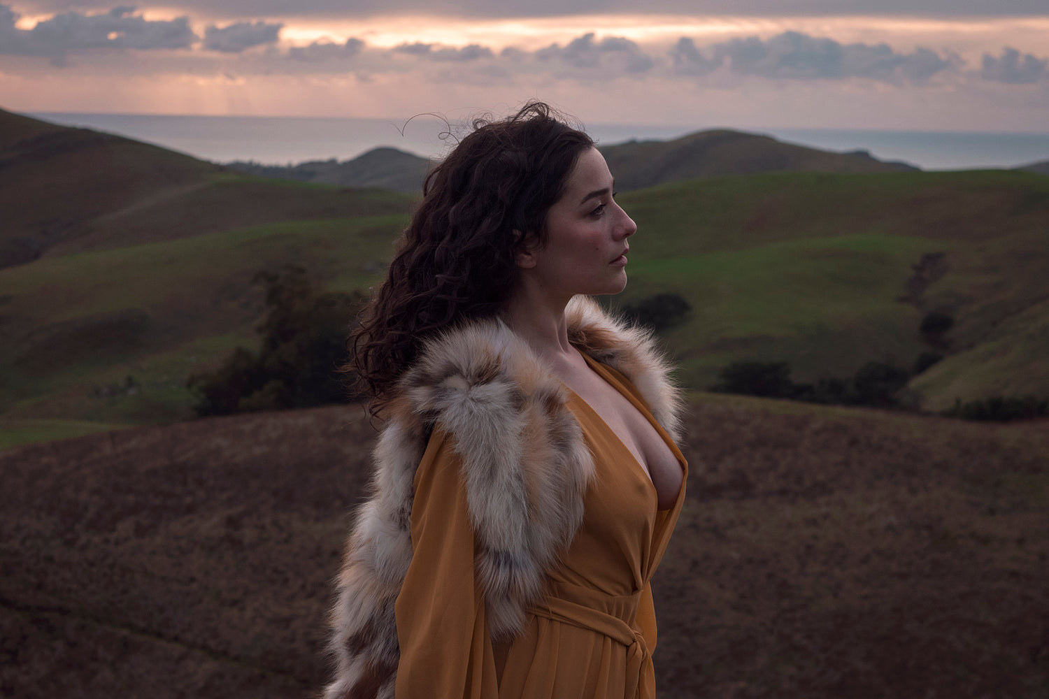 Xan from Stories of Evermore stands in front of a sprawling vista of green rolling hills and the distant ocean at sunset in a mustard yellow gown while wearing a vintage fur vest and vintage leather gloves.